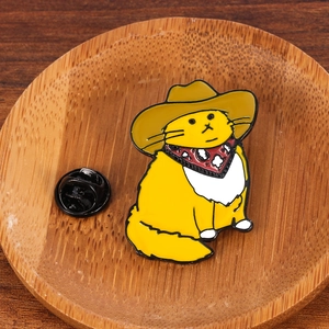 Detail of yellow cowboy cat enamel pin with a brown hat and red scarf on a wood tray