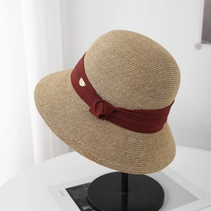 Détail d'un chapeau de paille beige avec ruban rouge foncé, nœud et logo doré sur un support