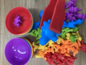 Toddler sorting toy detail showing a red tweezer picking a blue dinosaur over colorful bowls