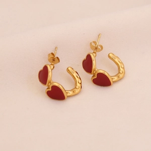 Detail of C-shaped golden earrings with double red heart charms on a soft surface