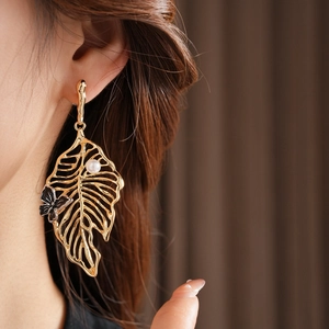 Close-up of golden hollow leaf earrings with a black butterfly and white pearl detail