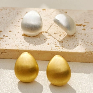 Brushed metal teardrop stud earrings in gold and silver finish on stone background