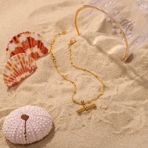 Detailed close-up of 18K gold-plated T-bar pendant on a fine anklet over beach sand