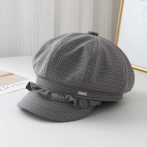 Detail of grey plaid newsboy cap featuring ruffle trim and a metal logo plaque