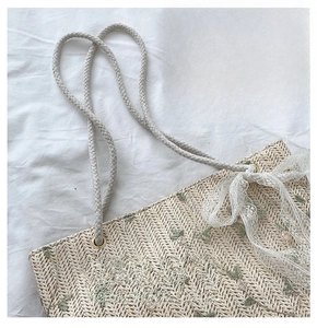 Detail of straw tote bag showing floral embroidery, lace bow, and braided shoulder strap