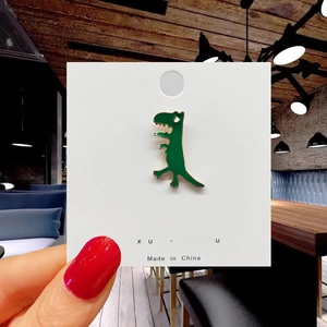 Detail of cute green dinosaur enamel pin on display card held against a blurred background