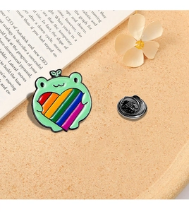 Close-up of a cute cartoon frog enamel pin holding a rainbow heart on an open book