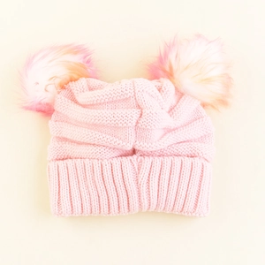 Pink knit children's winter hat with two colorful faux fur pompoms on plain background