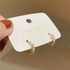 Detailed close-up of small gold pearl hoop earrings held on a Reina brand display card