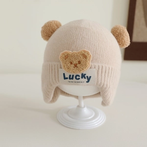 Detail of beige knit baby hat with twin pom-poms and plush bear patch for winter warmth