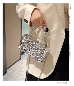 Silver textured box clutch bag with metal chain strap held by a model in a beige suit