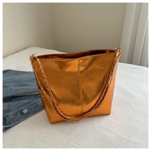 Close-up of glossy orange bucket bag with gold chain strap on soft white fabric