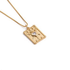 Détail du pendentif doré style tarot avec pierre en cœur et épées sur fond blanc