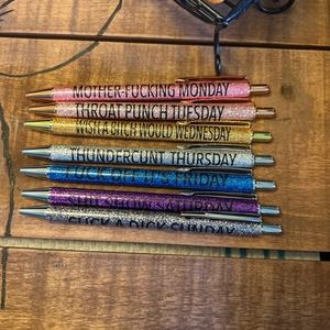 Detailed close-up of 7-pack glitter pens featuring profane weekly slogans on a wooden desk