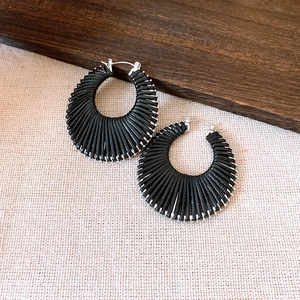 Black fan-shaped woven earrings with silver hoop fasteners on beige fabric surface