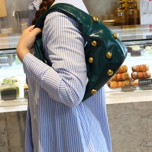 Dark green leather shoulder bag with gold rivets shown in a lifestyle bakery setting