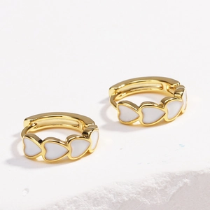 Detail shot of gold hoop earrings with white enamel heart charms on a textured background