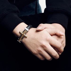 Detail of men's black braided leather bracelet featuring a golden steel cross on wrist