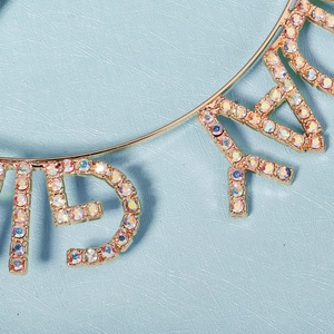 Sparkling rhinestone birthday crown with gold alloy letters on a light blue background