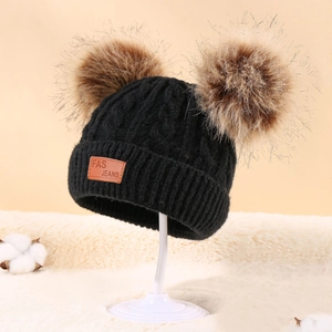 Detail of a black knit baby hat with two brown pom-poms and leather logo patch