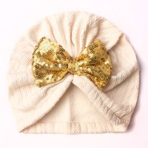 Detail of beige knitted baby hat featuring a large shimmering gold sequin bow accent