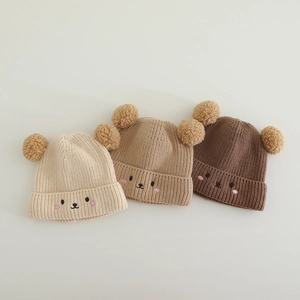 Detailed view of knitted baby hats with bear embroidery and double faux fur pom-poms