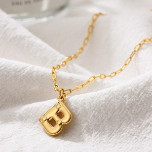Detail of a 3D bubble letter B pendant in gold on a white textured linen background