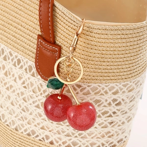 Sparkly red cherry keychain with gold hardware hanging on a woven straw bag