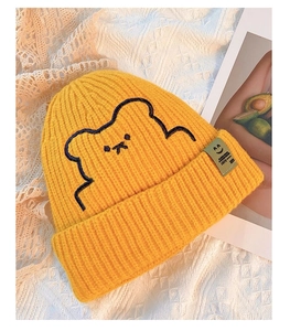 Close-up of yellow knitted beanie featuring cute bear embroidery and a green label