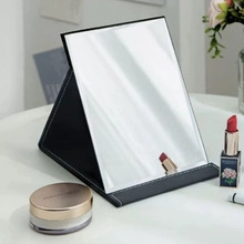 Close-up of a foldable portable vanity mirror with a black leather stand on a desk