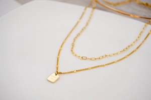 Close-up of double layer gold necklace featuring a Good Luck square pendant and paperclip chain