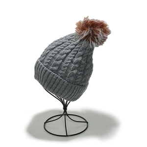 Gray knitted beanie with white fox fur pom-pom shown on a black metal display stand