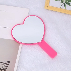 Pink heart-shaped handheld makeup mirror with handle placed on a soft plush white surface