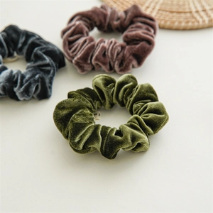 Close-up of three soft velvet scrunchies in dark green, grey, and brown on a beige table