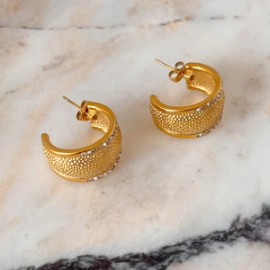 Golden C-shaped hoop earrings with hammered texture and zircons on marble surface