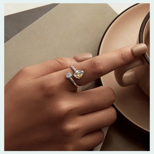 Detail of adjustable silver ring featuring a clear heart gem and gold flower on a finger