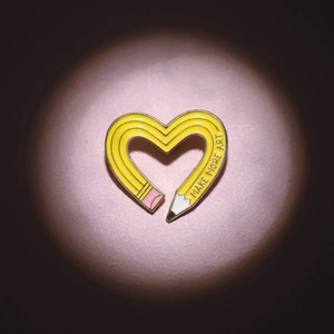 Detailed shot of yellow heart-shaped pencil enamel pin with 'MAKE MORE ART' text