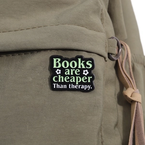 Macro shot of 'Books are cheaper than therapy' enamel pin on a khaki backpack