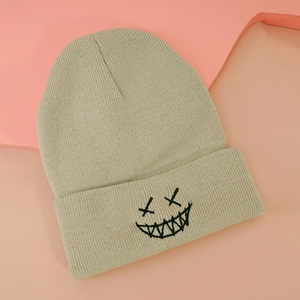 Close-up of beige ribbed knit beanie featuring black embroidered smiley face on folded brim