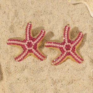 Exaggerated gold starfish earrings with pink and red rhinestones on a sandy beach