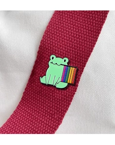 Close-up of a cute cartoon frog enamel pin with pride rainbow bar on a red bag strap