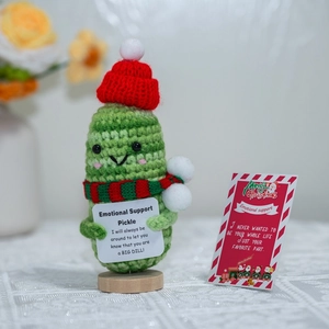 Close-up of handmade crochet Christmas pickle doll with holiday card on wooden base