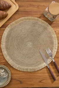Macro detail of round woven placemat with golden tassels on a rustic wooden dining table