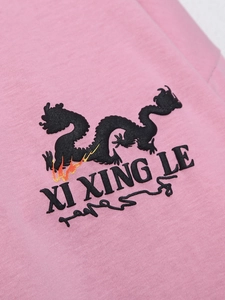 Nahaufnahme der schwarzen Drachenstickerei und des XI XING LE Textes auf rosa T-Shirt