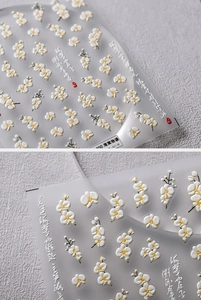 Dettaglio macro di adesivi 5D per unghie con fiori bianchi e gialli e decori calligrafici