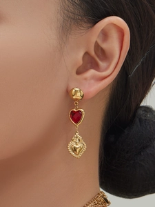 Close-up of heart-shaped gold earrings with red gemstone and sacred heart relief on model