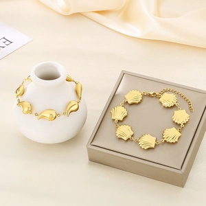 Golden stainless steel bracelet with shell and teardrop charms on a white vase and box