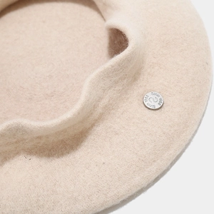 Detail of oat beige wool beret with circular silver metal logo plate and fine fabric texture