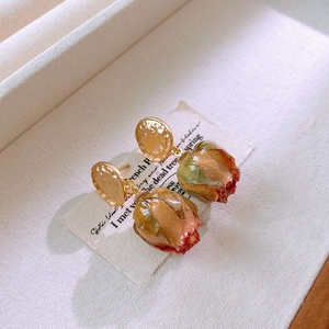 Macro shot of dried red rose bud earrings with textured golden metal studs on beige fabric