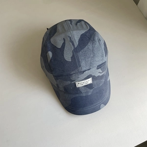 Detail of the 5-panel camouflage camp cap featuring a woven letter logo patch on the front